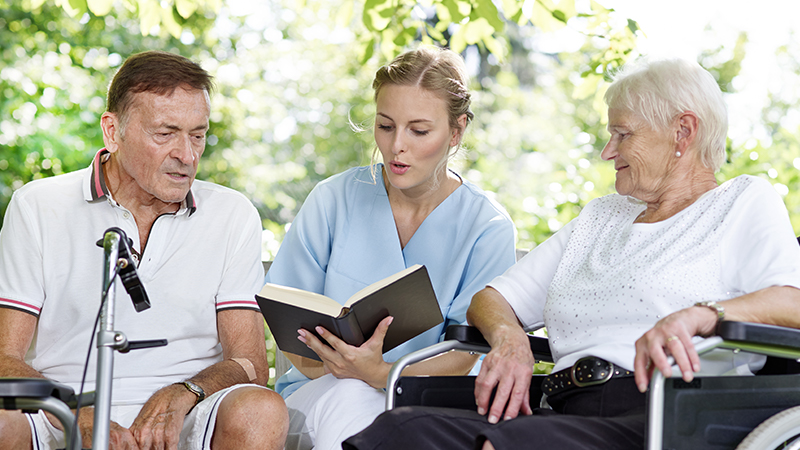 Junge Pflegerin, in der Mitte sitzend, liest 2 ältereren Personen etwas vor.