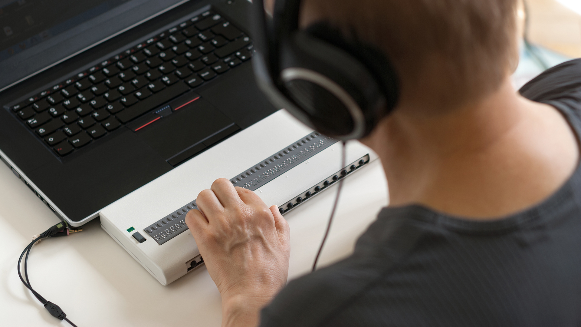 Person mit Kopfhörer an Braille-Tatstur vor Laptop.