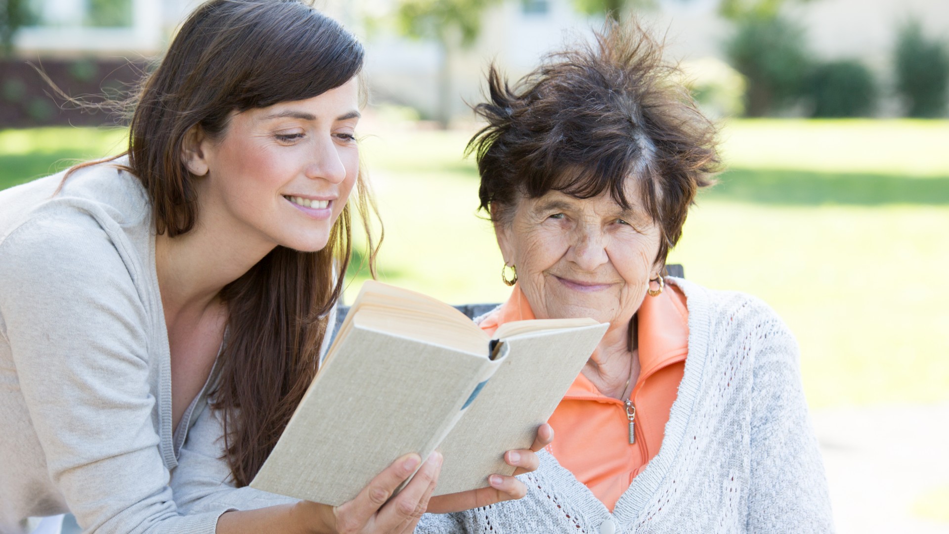 Jüngere Frau liest älterer Frau etwas aus einem Buch vor.